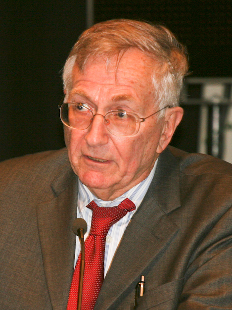 Seymour Hersh speaking at a conference table into a slim microphone, thick-framed glasses and red polka-dot tie under a grey suit, 2015