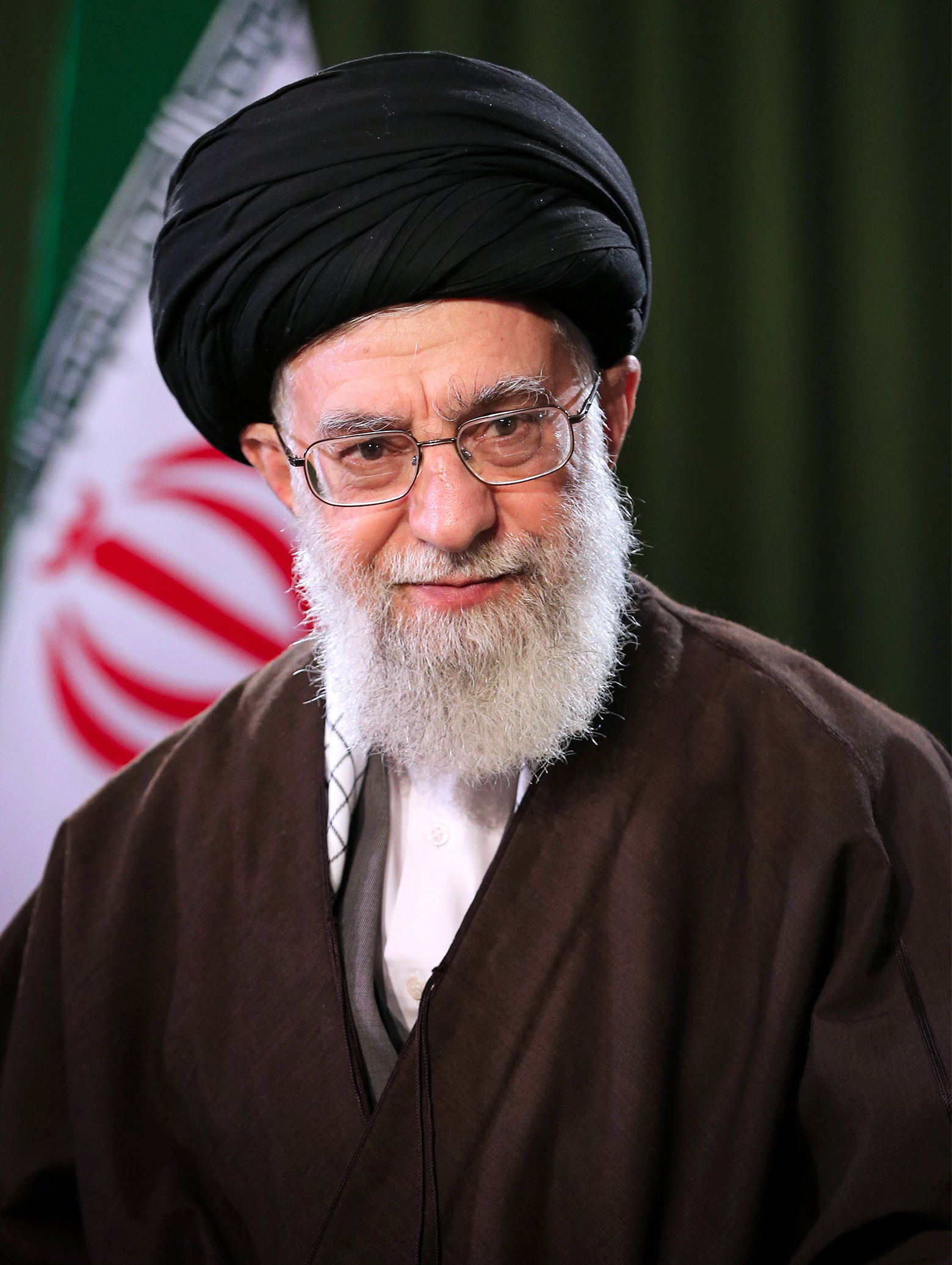 Ali Khamenei smiling in a black turban and brown robe over a white shirt, the Iranian flag behind him, 2018