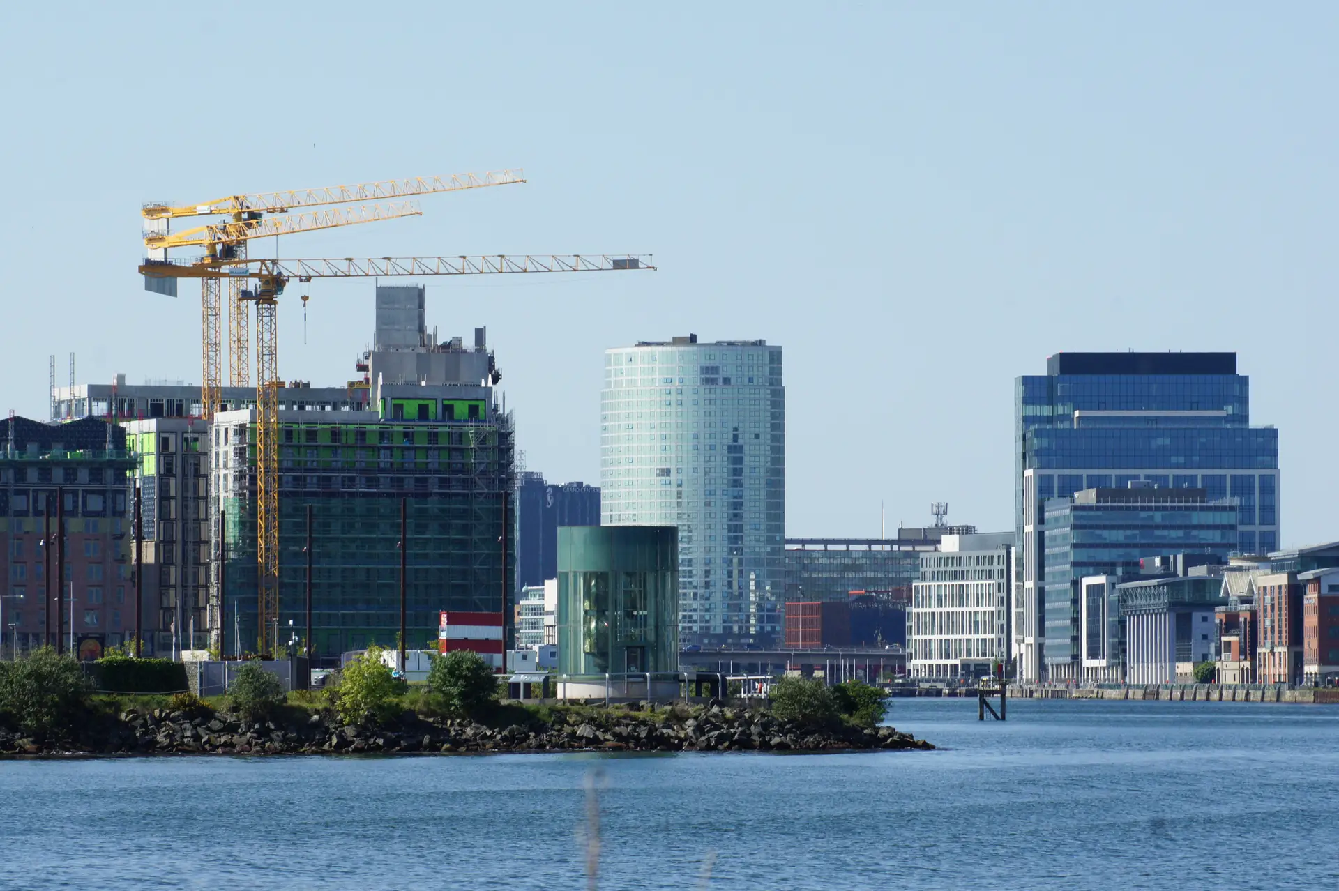 The Belfast waterfront under bright summer sky, tower cranes over new-build apartments beside glass office blocks along the River Lagan, 2015