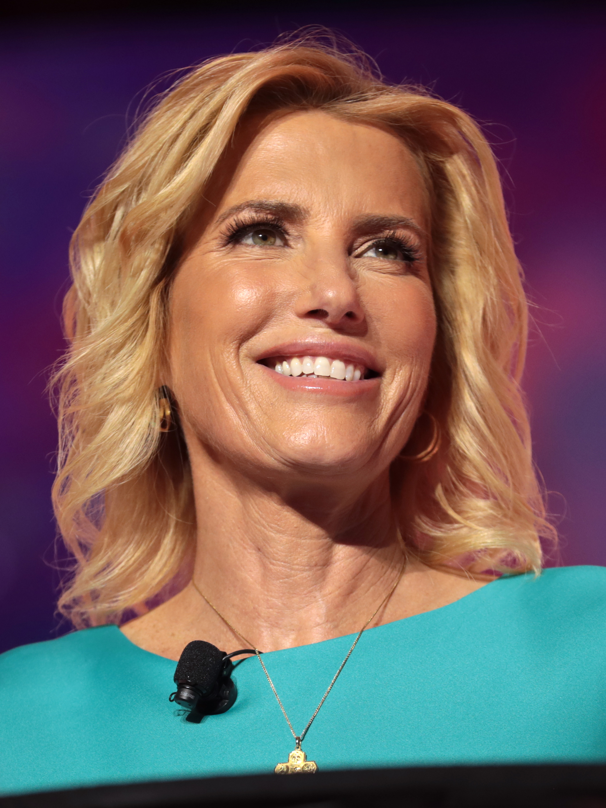 Laura Ingraham smiling on a CPAC stage in a teal dress with a lavaliere microphone clipped at the neck, 2018