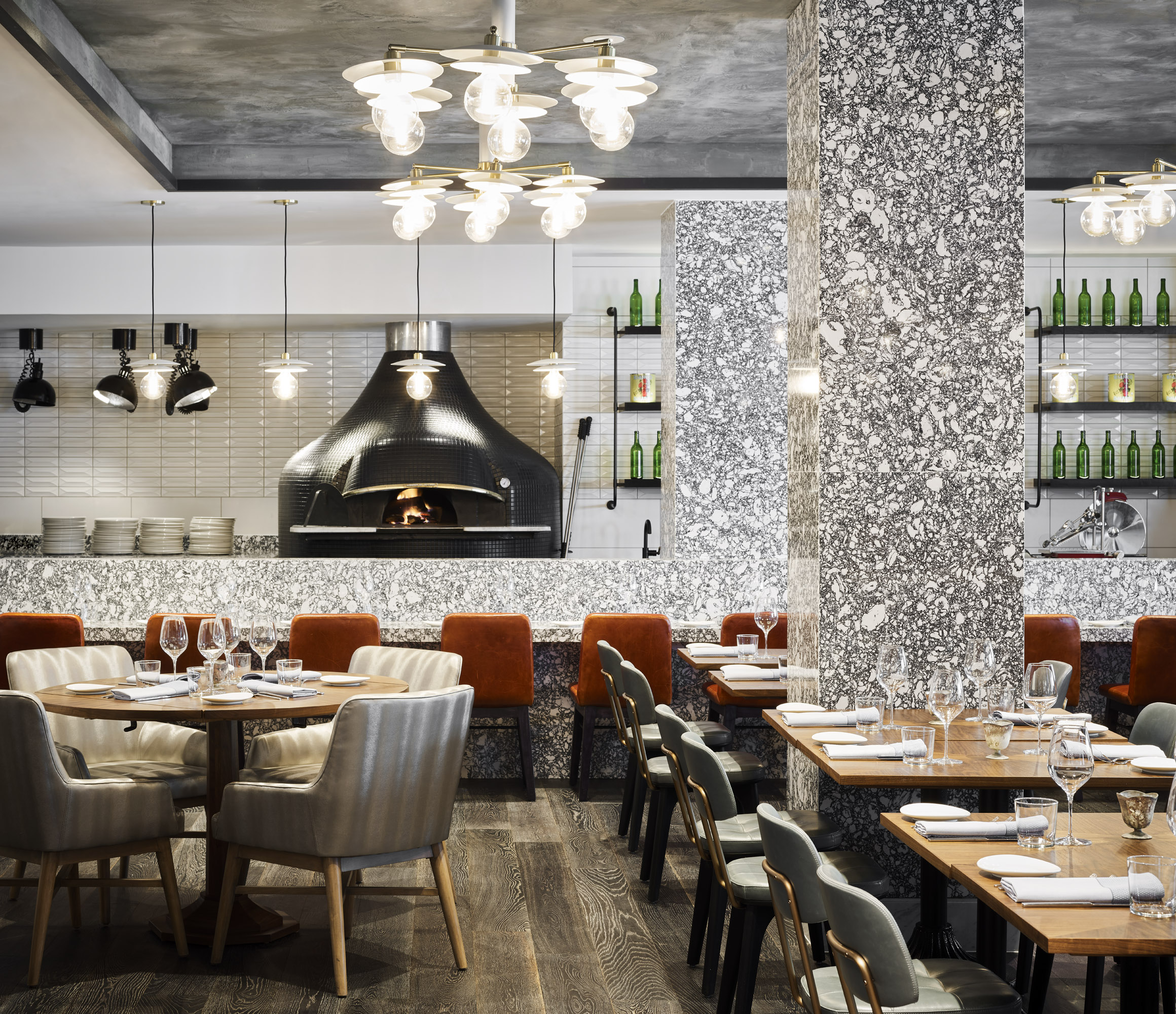 An empty restaurant dining room with a wood-fired pizza oven at the back, brass pendant lights over tables set with wine glasses, 2015