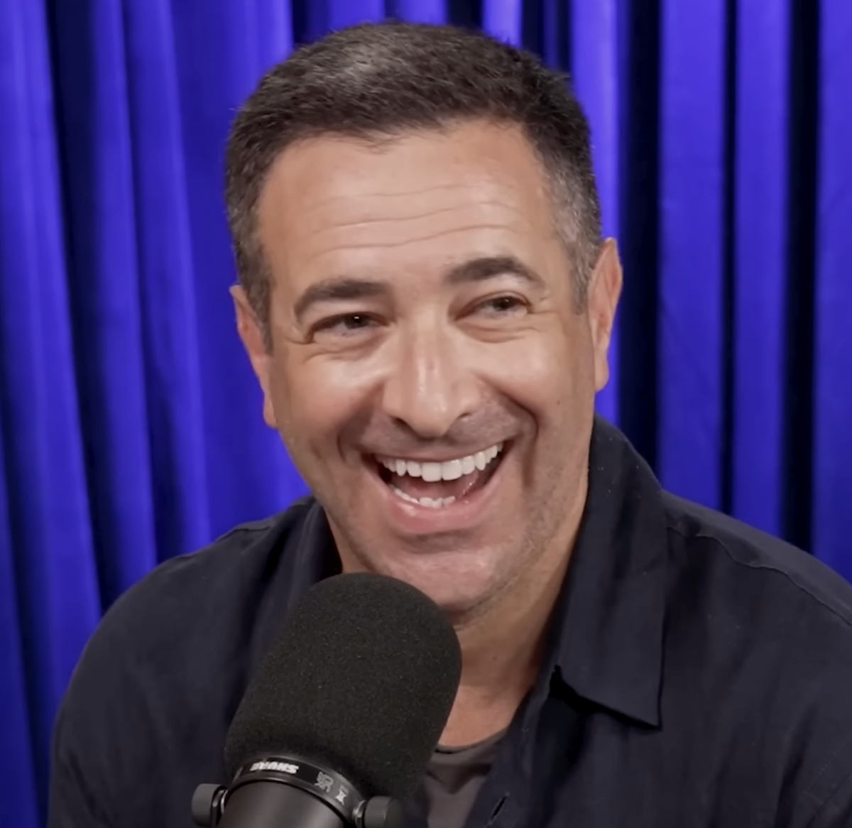 Ari Melber laughing at a podcast microphone in a black button-down against a blue curtain, 2019