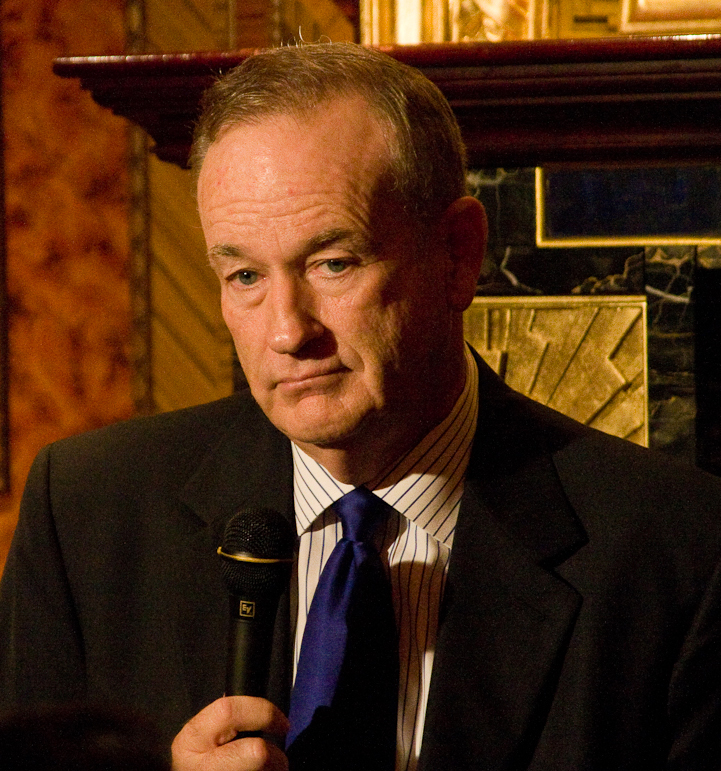 Bill O'Reilly holding a microphone in a dark blazer and blue tie before a fireplace mantel, 2010
