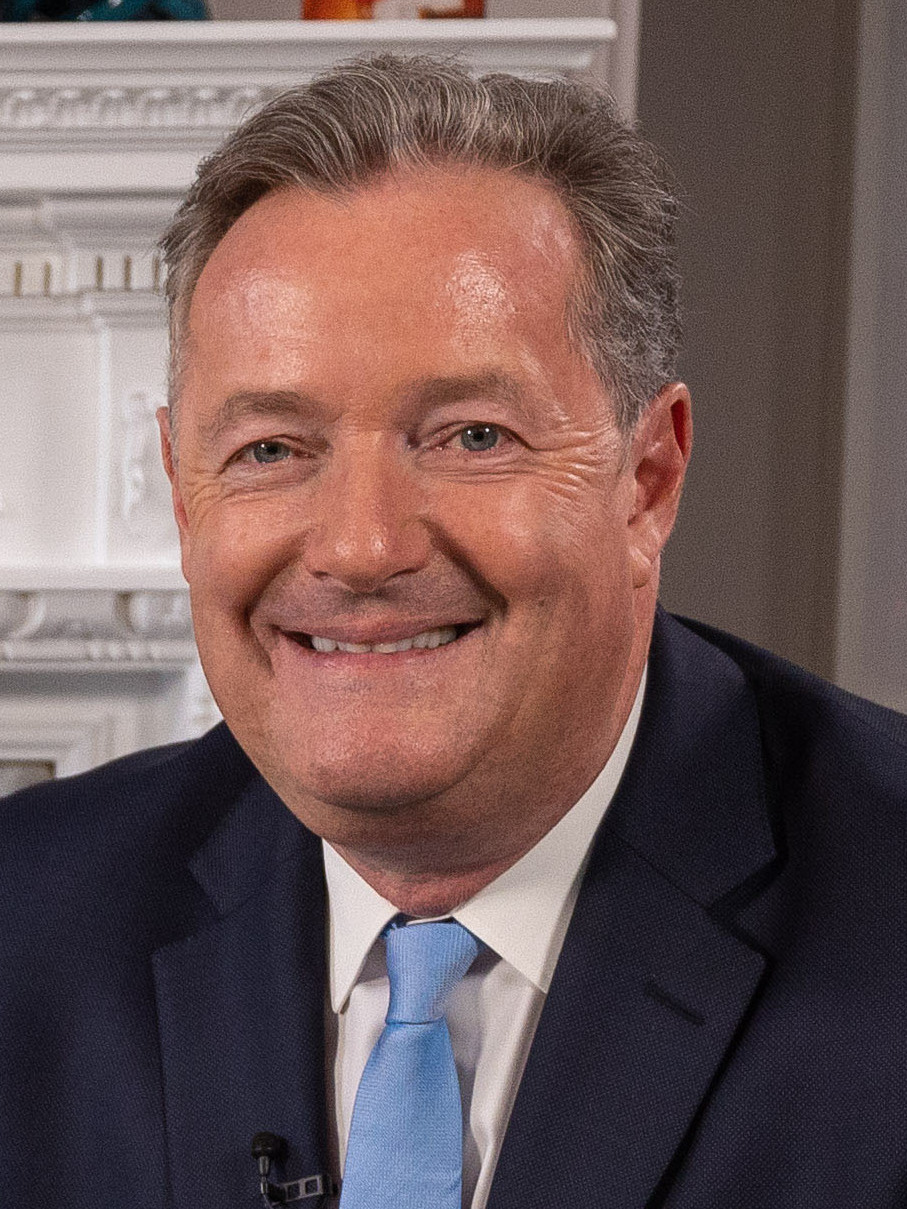 Piers Morgan smiling in a navy suit and light-blue tie before a white plaster mantelpiece, 2019
