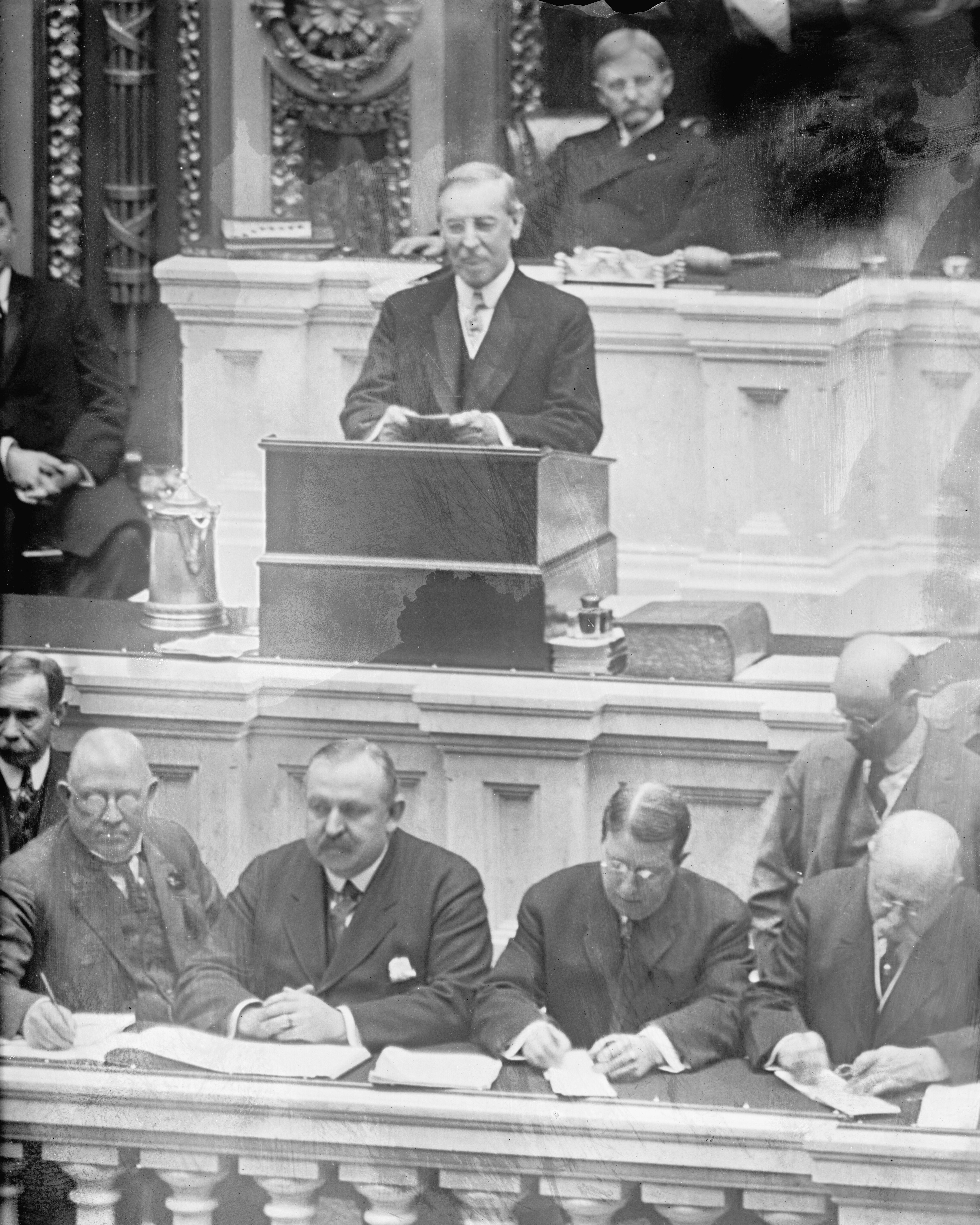 Woodrow Wilson at the rostrum before a joint session of Congress in a black-and-white photograph, aides taking notes at the desk below, 1917