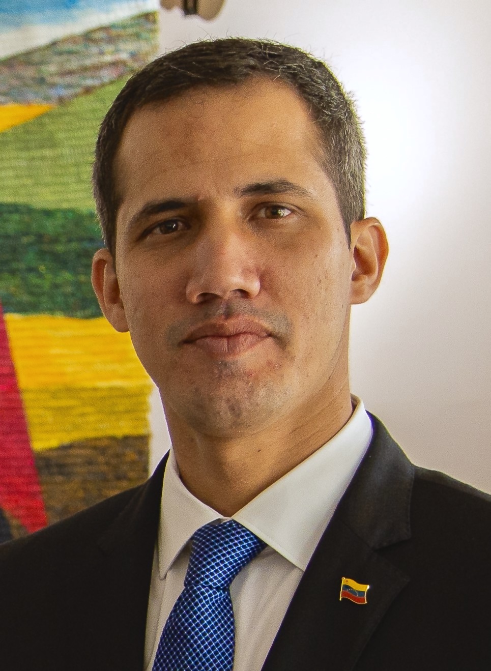 Juan Guaidó in a dark suit with a Venezuelan flag lapel pin, a landscape painting in yellow, blue and red on the wall behind him, 2019