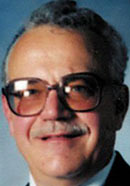 Gust Avrakotos in a dark jacket and moustache smiling into the lens of an old passport-style photograph, 1985