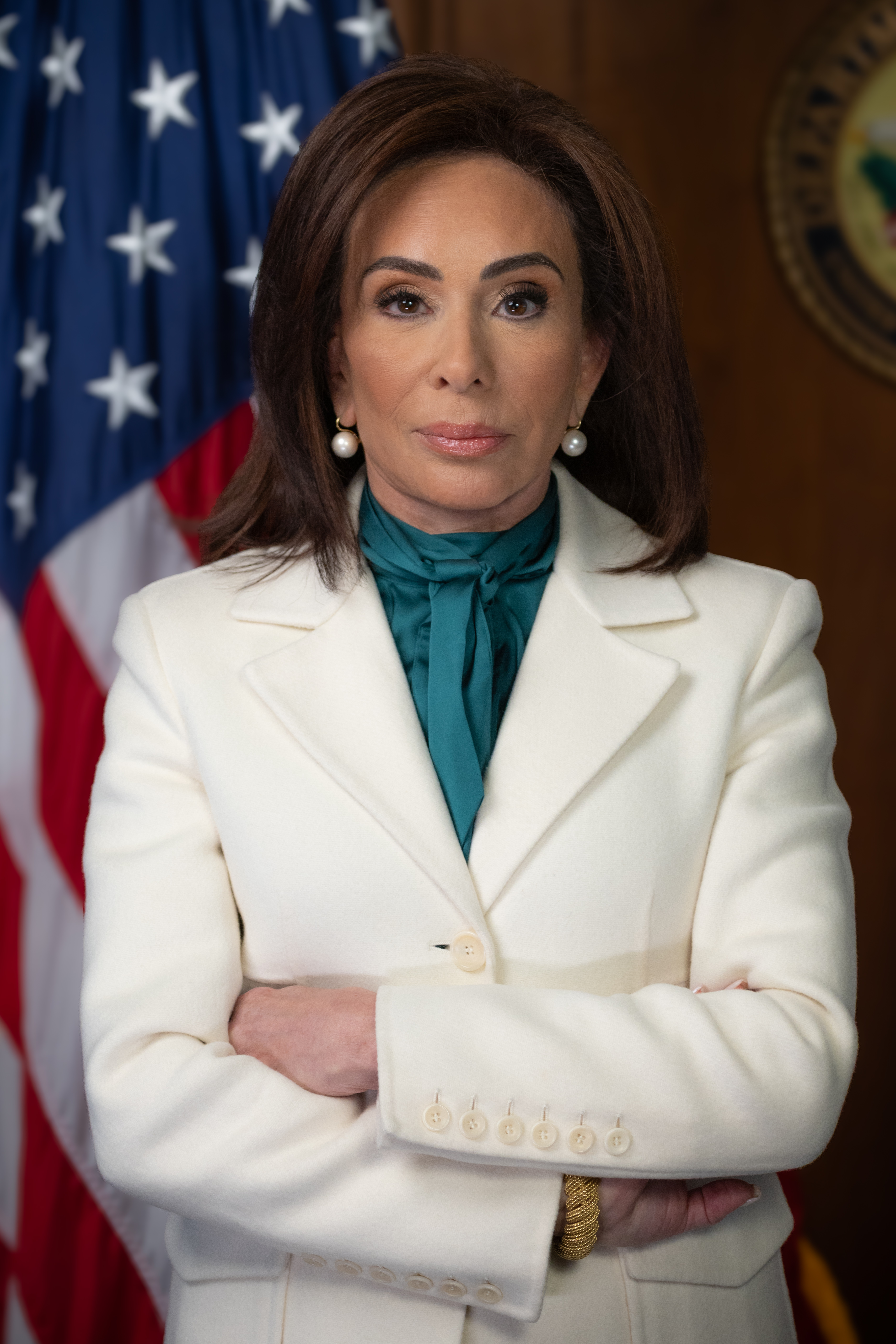 Jeanine Pirro in an official portrait, arms folded over a white blazer and teal pussy-bow blouse against a US flag and gold seal, 2025