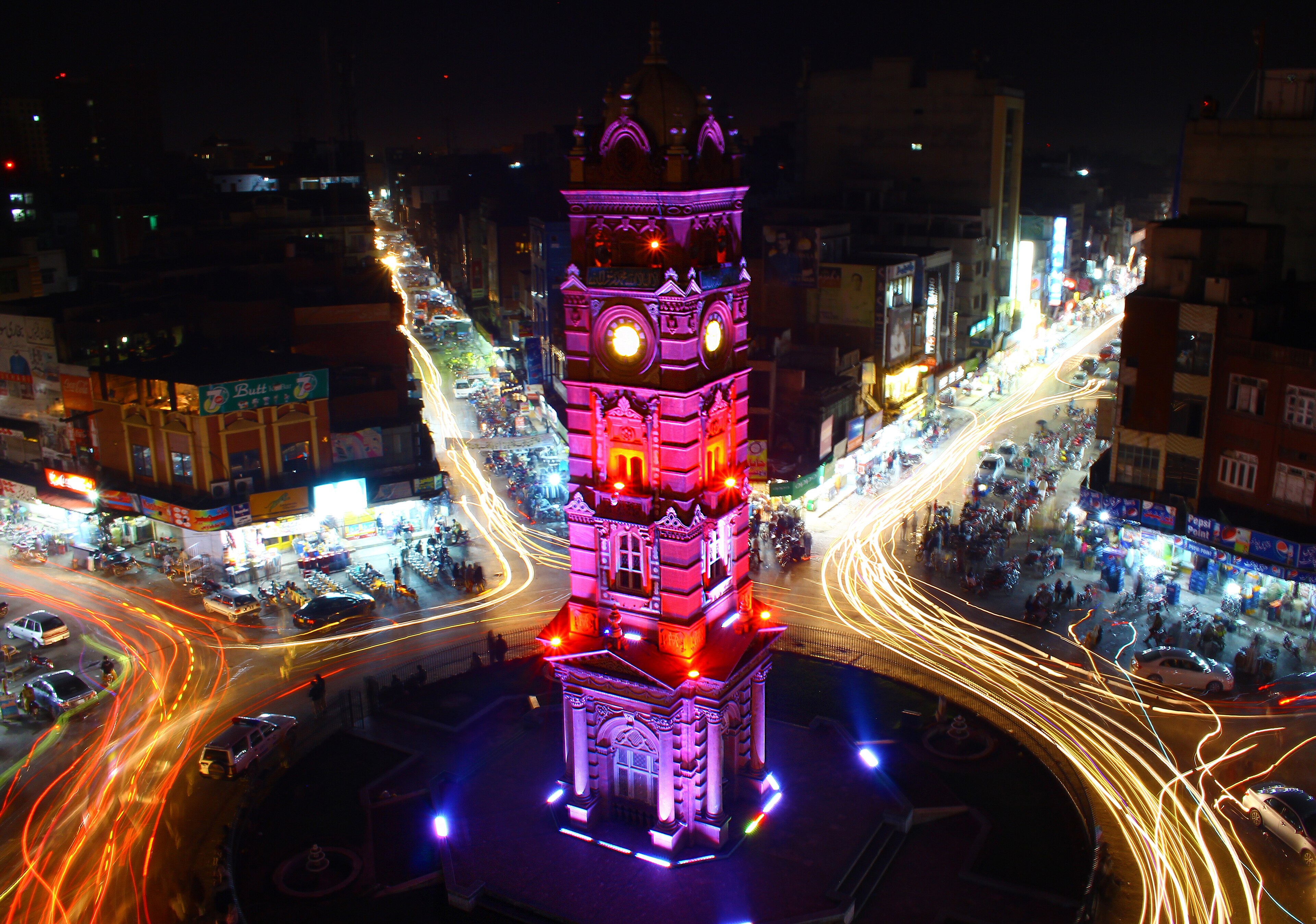 The Faisalabad Clock Tower lit magenta at night, traffic light trails streaming from a busy roundabout, 2015
