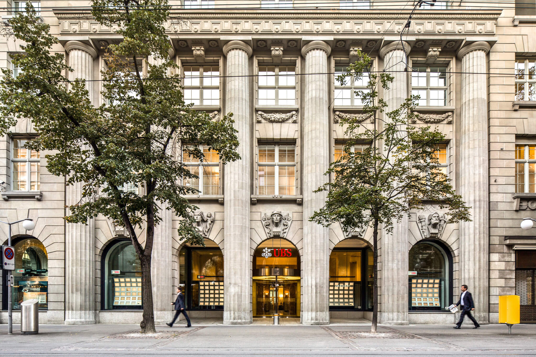 The colonnaded stone façade of the UBS headquarters on Bahnhofstrasse in Zurich, two suited pedestrians passing the entrance, 2016