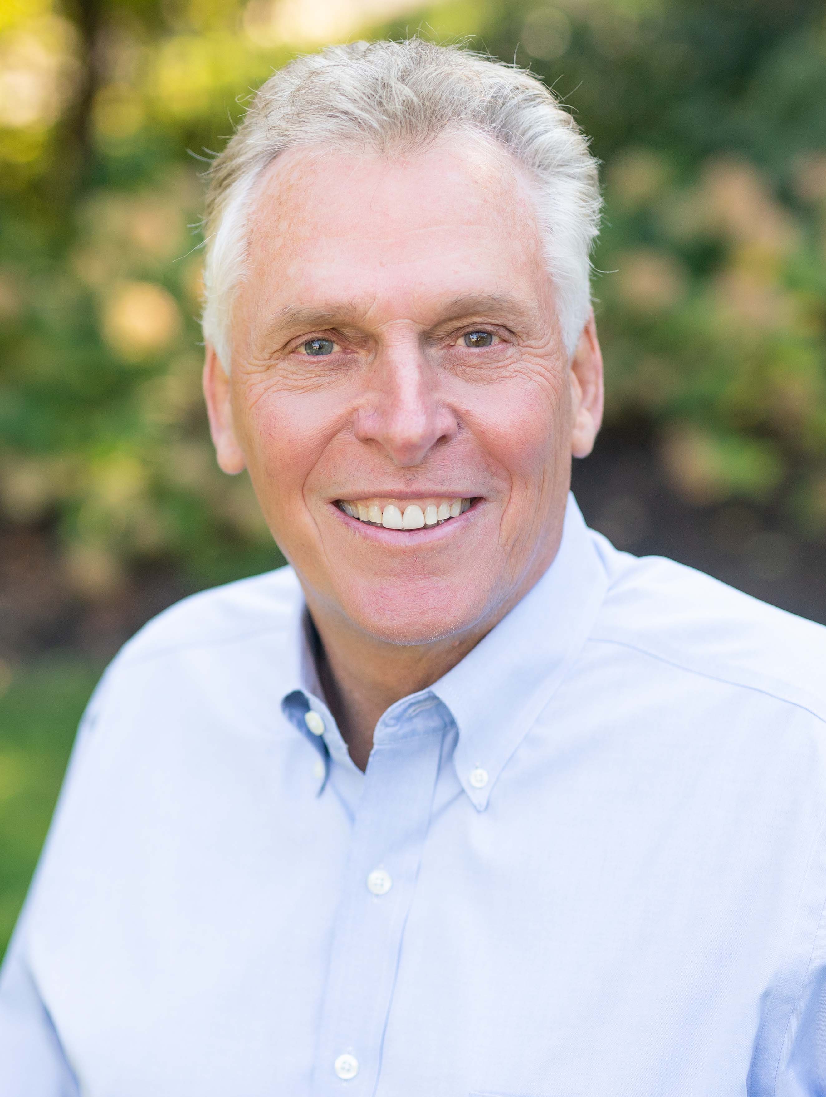 Virginia governor Terry McAuliffe in a light-blue button-down and no tie, smiling for a portrait against a leafy green backdrop, 2014