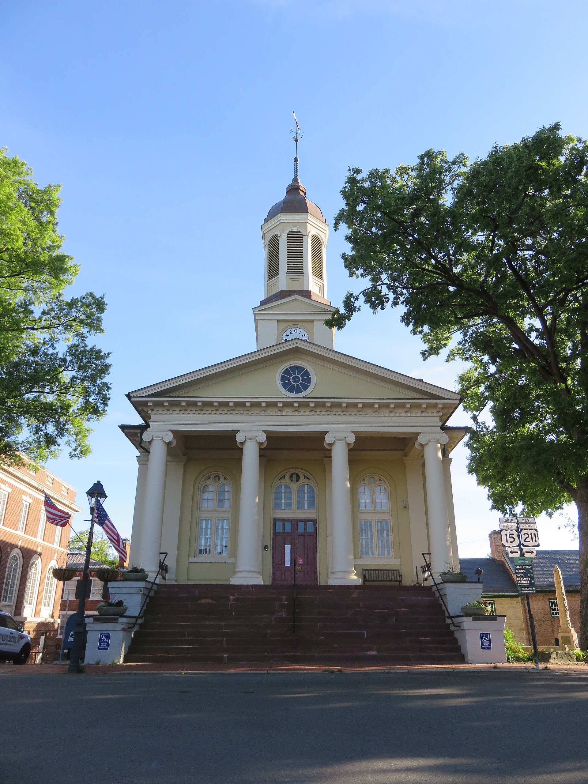 The pale-yellow Old Warrenton Courthouse in Virginia, a cupola and weathervane rising above its Ionic portico between two shading trees, 2015
