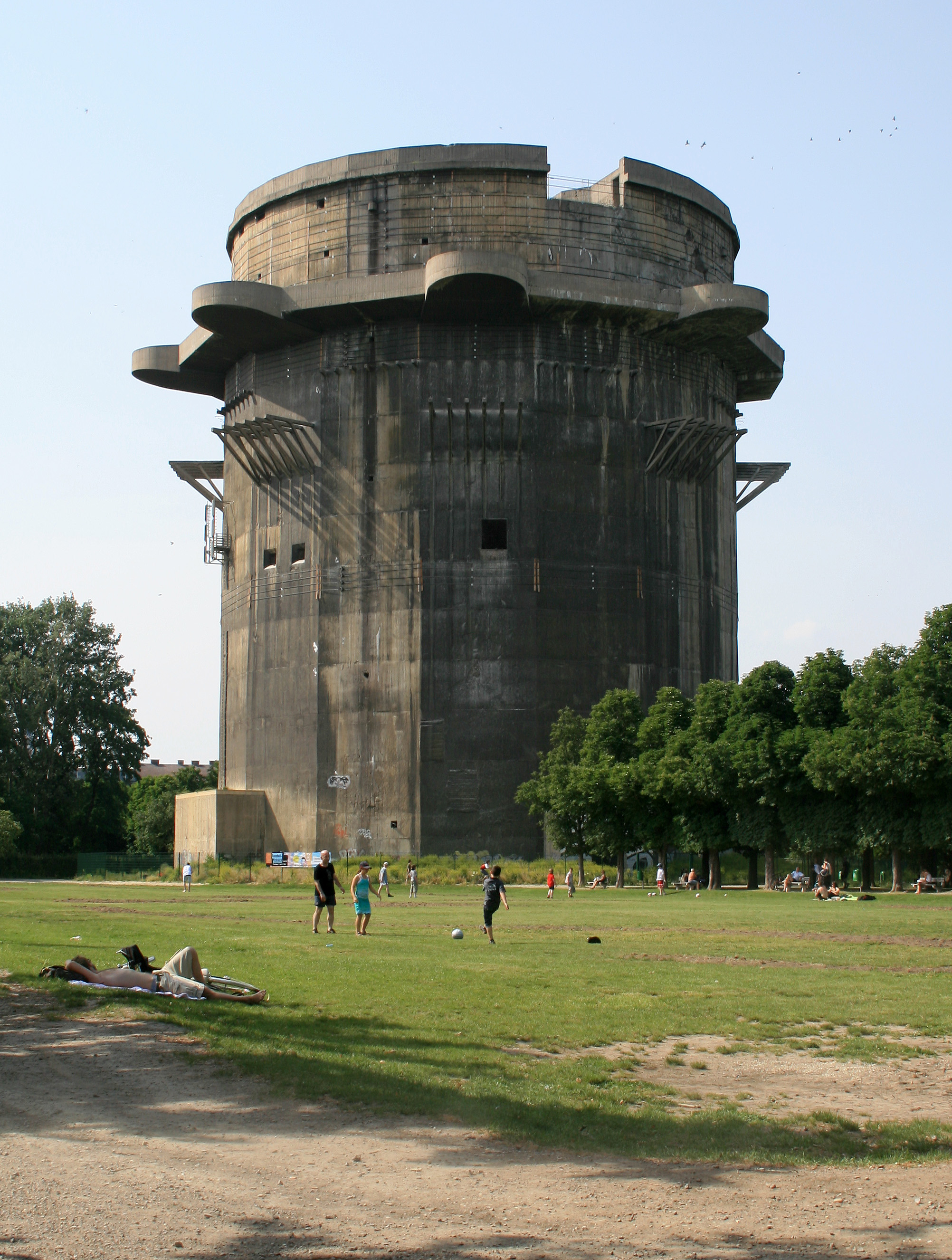 A massive concrete WWII flak tower standing over a park in Vienna, families picnicking on the grass at its foot, 2010