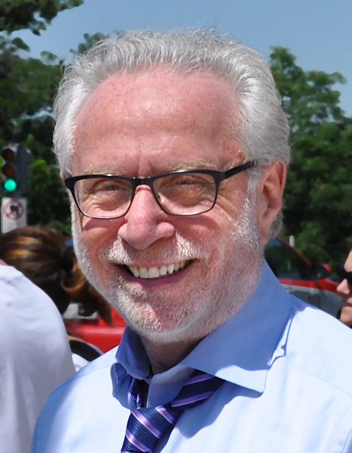 Wolf Blitzer in dark-framed glasses and a purple-striped tie smiling before a summer street backdrop of green trees, 2012
